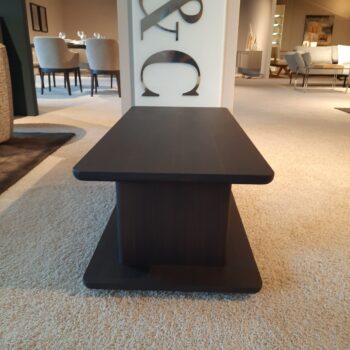 Rechteckiger Couchtisch Hugo Beistelltisch aus dunklem Holz mit zentraler Stütze, platziert auf einem beigen Teppich in einem modernen, gut beleuchteten Showroom von Molteni&C. Im Hintergrund sind Ess- und Loungemöbel zu sehen.