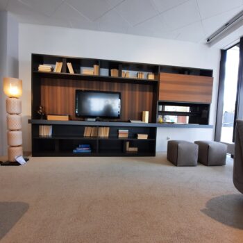 Ein modernes Wohnzimmer mit einem Molteni&C 505 Wohnwand-Bücherregal, einem Fernseher, Büchern, Dekorationsartikeln, einer einzigartigen Lampe, zwei Ottomanen und großen Fenstern, die natürliches Licht hereinlassen.
