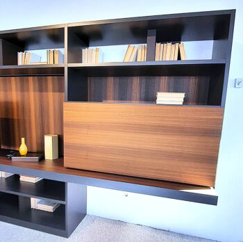 Ein modernes Bücherregal aus Holz, inspiriert vom Design von Molteni&C, mit Regalen, die teilweise mit gestapelten Büchern gefüllt sind, und zwei Dekorationsgegenständen – einer gelben Vase und einer rechteckigen Schachtel – auf einem unteren Regal, verleiht jedem Raum einen Wohnwand-Touch.
