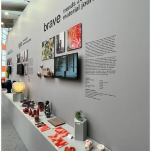 Ausstellungsdisplay mit an der Wand befestigten Texten, Fotos und einem Bildschirm über einem langen Tisch, auf dem verschiedene Keramik- und Designobjekte ausgestellt sind. Inspiriert von der Ambiente Frankfurt, zeigt die Wand mutige Trends 2021: Material Journey.
