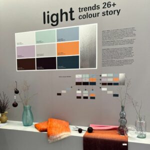 Eine Schautafel mit dem Titel light trends 26+ colour story, inspiriert von den Wohntrends 2026 auf der Ambiente Frankfurt, zeigt Farbmuster und Beschreibungen über einem Regal mit Vasen, getrockneten Pflanzen, Glaswaren und gefalteten orangefarbenen und braunen Textilien.