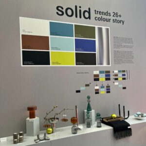 Eine Ausstellungswand auf der Ambiente Frankfurt zeigt eine Farbpalettenkarte mit dem Titel solid trends 26+ colour story, auf der Objekte in verschiedenen Formen und Farben in einem Regal angeordnet sind - ein Blick auf die Wohntrends 2026.