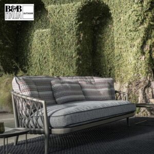 Ein modernes Outdoor-Sofa mit gemusterten Kissen steht auf einem dunklen Teppich vor efeubewachsenen Wänden - perfekt für den Frühling und den Outdoor-Saisonstart. B&B Italia Outdoor-Logo in der Ecke. Entdecken Sie mehr bei Möbel Braum.
