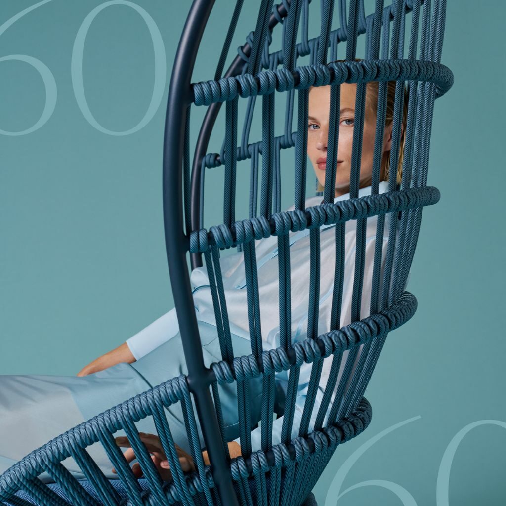 Eine Person sitzt in einem modernen blauen geflochtenen Kettal-Stuhl, der teilweise von der ikonischen Struktur des Stuhls verdeckt wird, vor einem blaugrünen Hintergrund, auf dem die Zahl 60 schwach zu erkennen ist.