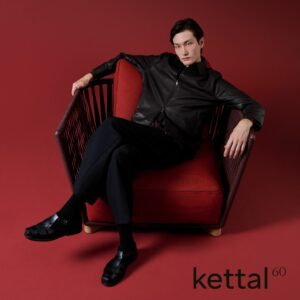Ein Mann in dunkler Kleidung sitzt entspannt in einem modernen roten Sessel auf rotem Boden. Das Wort Kettal, bekannt für ikonische Outdoormöbel, und die Zahl 60 erscheinen in der unteren rechten Ecke.