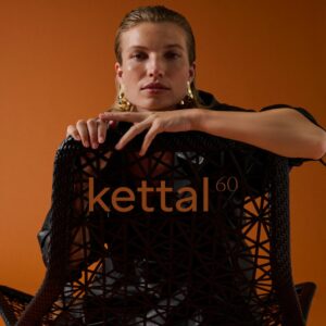 Eine Person mit zurückgekämmtem Haar sitzt hinter einem geflochtenen Kettal-Stuhl mit Kettal 60 auf der Rückenlehne, der ikonische Outdoormöbel vor einem orangefarbenen Hintergrund zeigt.