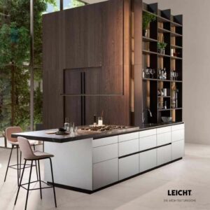 Moderne Küche mit einer großen Insel, integriertem Kochfeld, offenen Regalen und Barhockern. Mit Holzakzenten und bodentiefen Fenstern - ein Musterbeispiel für Interior Design 2026 von Braum Interior Design. LEICHT-Logo in der unteren rechten Ecke.