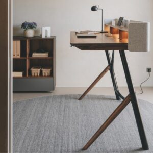 Ein moderner Schreibtisch mit angewinkelten Beinen steht auf einem grauen Teppich, perfekt für das Homeoffice. Eine kleine Lampe, Bücher und Papiere stehen auf dem Schreibtisch. Im Hintergrund sorgt ein Holzregal mit Ordnern und Körben für schön wohnen Charme.
