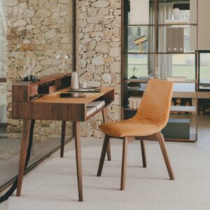 Ein moderner Holzschreibtisch mit einem orangefarbenen Polsterstuhl steht auf einem hellen Teppich in einem Homeoffice, in dem eine Steinmauer und ein großes Fenster einen inspirierenden Raum für besseres Arbeiten schaffen, während Regale im Hintergrund eine schöne Außenlandschaft einrahmen.