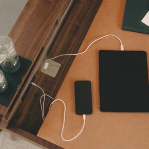 Ein Smartphone und ein Tablet werden auf einem Schreibtisch mit braunen Leder- und Holzoberflächen aufgeladen und bilden zusammen mit einem geschlossenen Notebook und einer gläsernen Sanduhr eine Homeoffice-Einrichtung, die zu schön wohnen und besser arbeiten inspiriert.