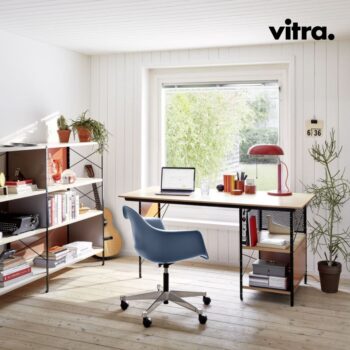 Ein minimalistisches Homeoffice mit einem eleganten Eames-Schreibtisch und einem blauen Stuhl, einem Laptop, einer roten Lampe, Bücherregalen, Pflanzen und einem großen Fenster, durch das Tageslicht hereinströmt. In der rechten oberen Ecke ist der Markenname „vitra.“ zu sehen.