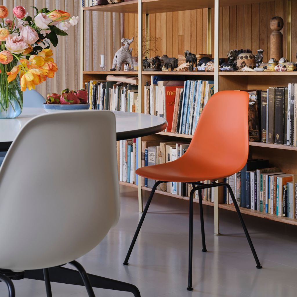 Ein modernes Zimmer mit einem runden Tisch, weißen und orangefarbenen Vitra Eames DSX Side Chairs, Bücherregalen voller Bücher und Figuren sowie einer Vase mit Blumen auf dem Tisch. Ein modernes Zimmer mit einem runden Tisch, weißen und orangefarbenen Vitra Eames DSX Side Chairs, Bücherregalen voller Bücher und Figuren sowie einer Vase mit Blumen auf dem Tisch.