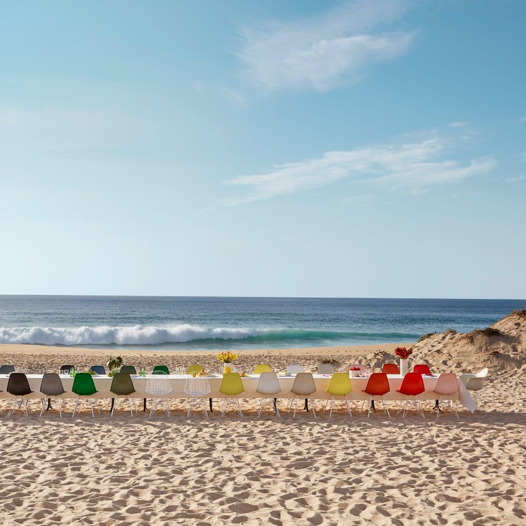 Eine Reihe bunter Eames Side Chairs steht an einem Sandstrand mit Blick auf das Meer unter einem strahlend blauen Himmel. Eine Reihe bunter Eames Side Chairs steht an einem Sandstrand mit Blick auf das Meer unter einem strahlend blauen Himmel.