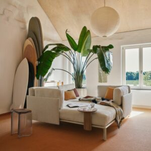 Ein modernes Wohnzimmer mit einem weißen Sektionssofa, einer großen Grünpflanze und Surfbrettern, die in der Ecke gestapelt sind, mit Vitra-Stücken, die Harmonie nach Hause bringen, wenn das Sonnenlicht durch das Fenster strömt.