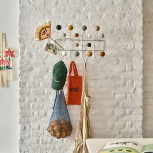 Bringen Sie die Harmonie des Winters nach Hause mit dieser Vitra Homestories Garderobe an einer weißen Backsteinwand, die eine grüne Mütze, eine Netztasche, ein gestreiftes Kleidungsstück, eine orangefarbene Tragetasche und ein Kinderkunstwerk präsentiert.