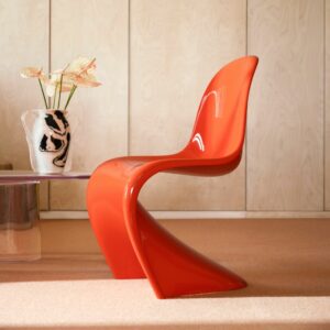 Ein moderner orangefarbener Kunststoffstuhl mit geschwungenem Design steht neben einem Glastisch und einer Blumenvase und bringt Vitra Homestories und Harmonie zu Hause in einen minimalistischen Raum mit holzgetäfelten Wänden.