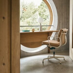 Ein moderner Vitra-Bürostuhl steht an einem Holzschreibtisch vor einem großen runden Fenster mit Blick ins Grüne; eine Lampe und Dekorationsartikel verleihen diesem einladenden Arbeitsbereich Harmonie zu Hause.