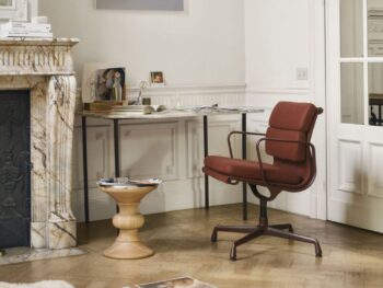 Ein modernes Home Office mit rotem Drehstuhl, hölzernem Beistelltisch und Marmorkamin strahlt Harmonie zu Hause aus. Aufgeschlagene Bücher und Zeitschriften, die auf dem Holzboden verstreut liegen, laden in dieser hellen Vitra Homestories Umgebung zur gemütlichen Winterinspiration ein.