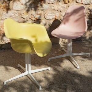Zwei moderne Stühle, ein gelber mit Armlehnen und ein blassrosa ohne, von Vitra, stehen auf einer Steinterrasse mit weißen Metallfüßen neben einer strukturierten Steinwand - ein Schaufenster für echte Designklassiker.