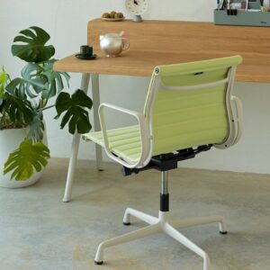 Ein hellgrüner Vitra-Bürostuhl mit weißem Gestell steht vor einem Holzschreibtisch mit Teeservice, Uhr und Pflanze auf einem Betonboden - ein subtiler Designklassiker für jeden Arbeitsbereich.