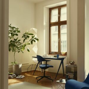 Ein helles Homeoffice mit einem großen Fenster, einem blauen Stuhl und Schreibtisch, einer hohen Pflanze, einer geometrischen Lampe und einem Bücherstapel auf dem Boden - perfekt zum schön wohnen und besser arbeiten.