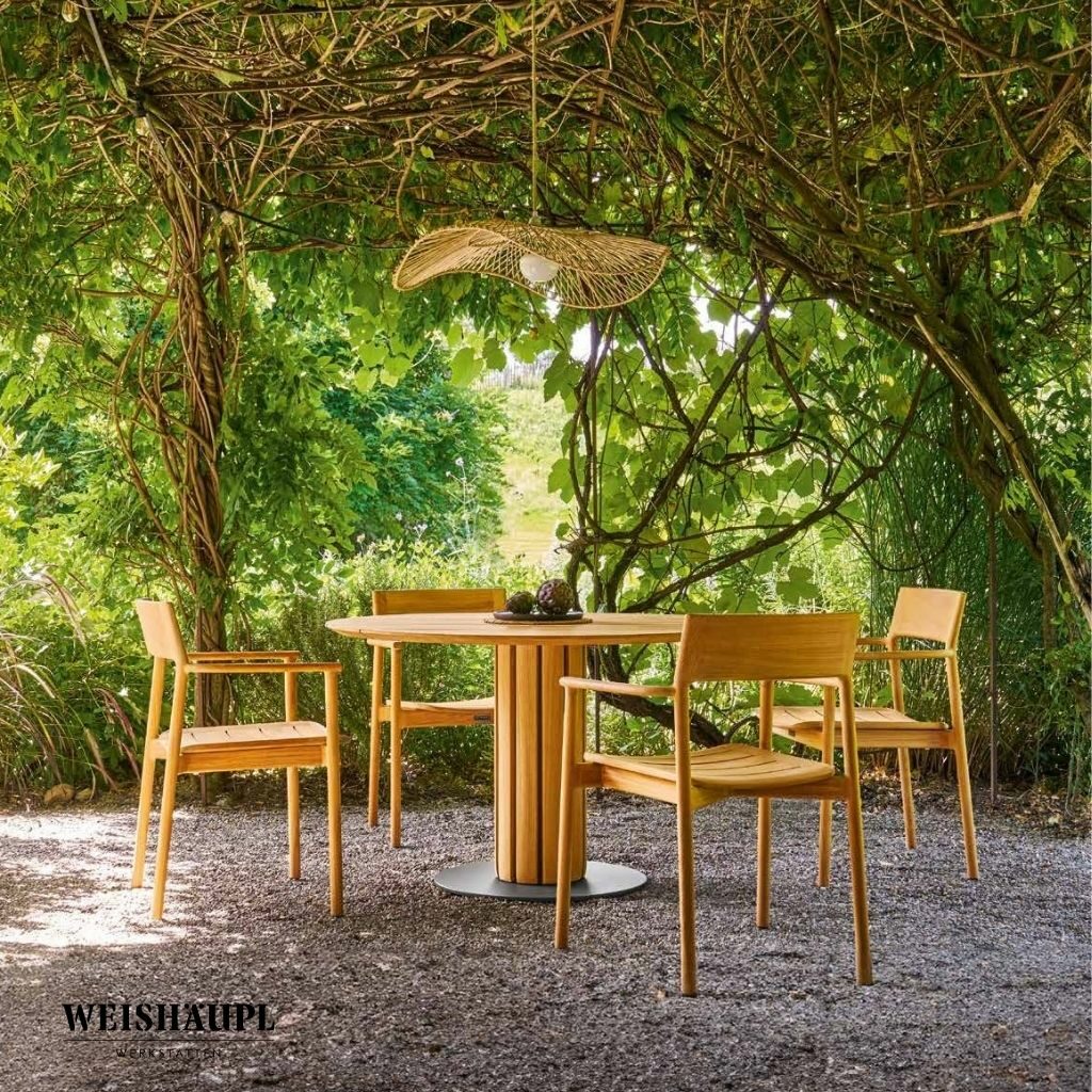 Vier Holzstühle und ein runder Tisch stehen unter einer mit Weinreben bewachsenen Pergola mit einer geflochtenen Leuchte. So entsteht ein einladender Sitzbereich im Freien, der den zeitlosen Touch von Braum Interior Design-Räume die wirken widerspiegelt.