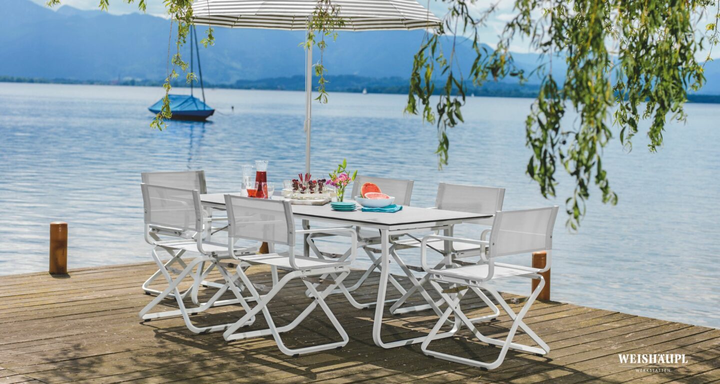 Weiße Design-Gartenmöbel mit sechs Stühlen und einem Sonnenschirm auf einem Holzsteg an einem See, mit Getränken und Snacks; Berge, ein Segelboot und die Qualität von Möbel Braum vervollständigen die Szene.