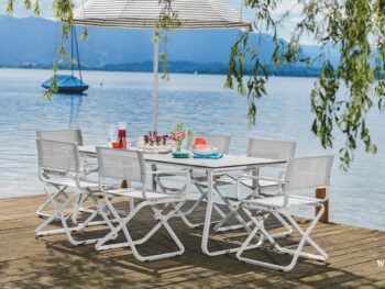 Weiße Design-Gartenmöbel mit sechs Stühlen und einem Sonnenschirm auf einem Holzsteg an einem See, mit Getränken und Snacks; Berge, ein Segelboot und die Qualität von Möbel Braum vervollständigen die Szene.