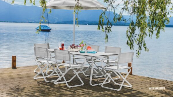 Weiße Design-Gartenmöbel mit sechs Stühlen und einem Sonnenschirm auf einem Holzsteg an einem See, mit Getränken und Snacks; Berge, ein Segelboot und die Qualität von Möbel Braum vervollständigen die Szene.