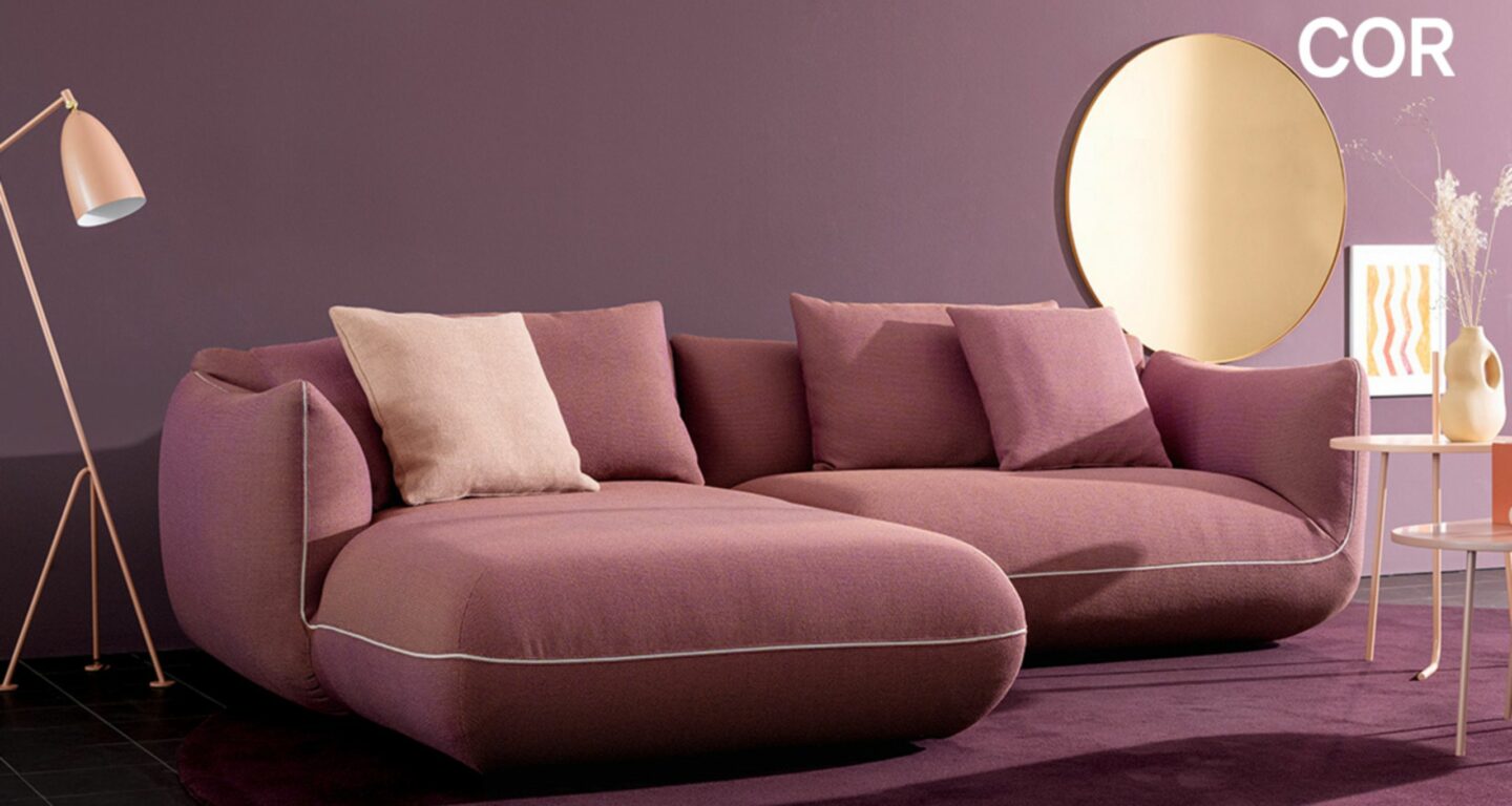 Ein modernes Wohnzimmer mit einem lilafarbenen Sektionssofa, passenden Kissen, einem runden Spiegel und einer rosafarbenen Stehlampe vor einer lilafarbenen Wand - perfekt für alle, die während der COR Vorteilswochen bei Möbel Braum nach Designklassikern suchen. Ein modernes Wohnzimmer mit einem lilafarbenen Sektionssofa, passenden Kissen, einem runden Spiegel und einer rosafarbenen Stehlampe vor einer lilafarbenen Wand - perfekt für alle, die während der COR Vorteilswochen bei Möbel Braum nach Designklassikern suchen.