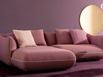 Ein modernes Wohnzimmer mit einem lilafarbenen Sektionssofa, passenden Kissen, einem runden Spiegel und einer rosafarbenen Stehlampe vor einer lilafarbenen Wand - perfekt für alle, die während der COR Vorteilswochen bei Möbel Braum nach Designklassikern suchen.