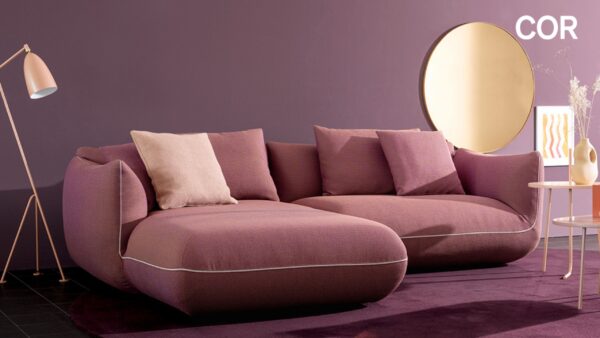 Ein modernes Wohnzimmer mit einem lilafarbenen Sektionssofa, passenden Kissen, einem runden Spiegel und einer rosafarbenen Stehlampe vor einer lilafarbenen Wand - perfekt für alle, die während der COR Vorteilswochen bei Möbel Braum nach Designklassikern suchen.