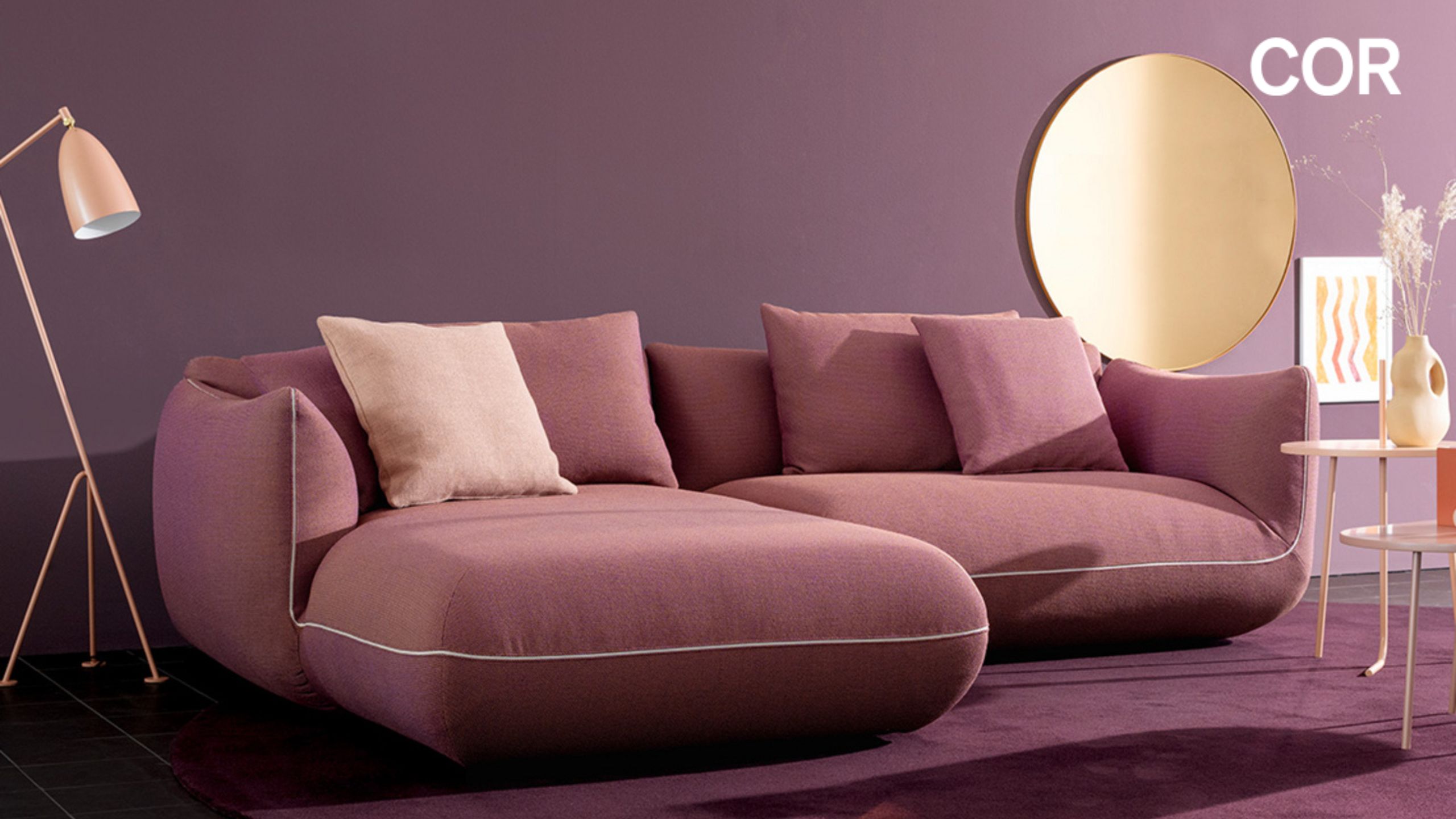 Ein modernes Wohnzimmer mit einem lilafarbenen Sektionssofa, passenden Kissen, einem runden Spiegel und einer rosafarbenen Stehlampe vor einer lilafarbenen Wand - perfekt für alle, die während der COR Vorteilswochen bei Möbel Braum nach Designklassikern suchen.