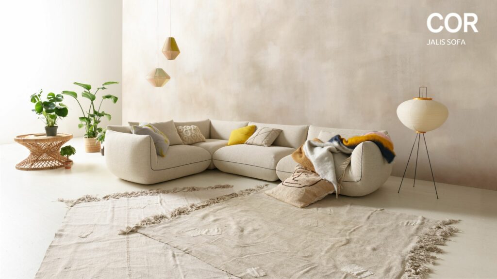 Ein modernes Wohnzimmer mit einem beigen Sektionssofa, einem neutralen Teppich, einer Stehlampe, einem kleinen Tisch mit einer Topfpflanze und geometrischen Pendelleuchten; COR JALIS SOFA Text in der Ecke - entdecken Sie Designklassiker bei Möbel Braum während der COR Vorteilswochen.
