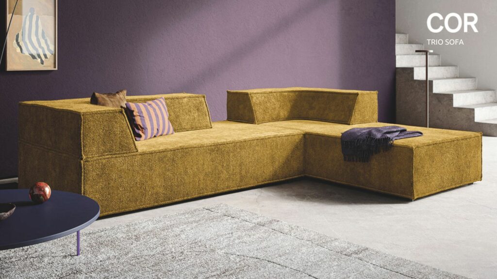 Modernes senfgelbes Sektionssofa mit geometrischem Design von Designklassiker, akzentuiert mit lilafarbenen Kissen und Überwurf, vor einer lilafarbenen Wand in der Nähe einer Treppe und eines runden Tisches - jetzt erhältlich bei Möbel Braum während der COR Vorteilswochen.