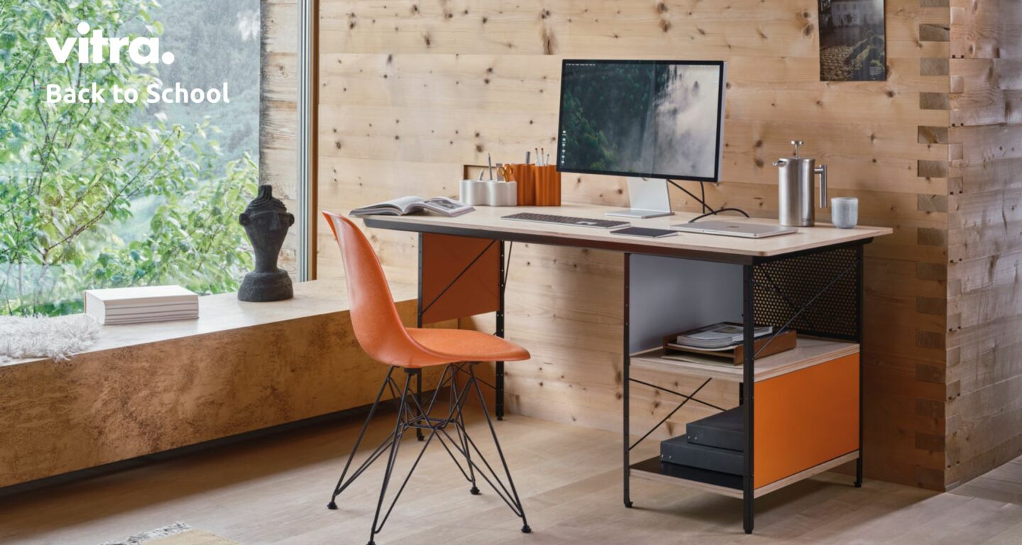 Ein modernes Heimbüro verfügt über einen Schreibtisch aus Holz mit orangefarbenen Akzenten, eine Eames-Schreibtischkombination, einen orangefarbenen „Back to School“-Stuhl von Vitra, einen Computermonitor, Bücher und Schreibwaren – vor dem Hintergrund einer holzvertäfelten Wand mit einem Fenster, das den Blick auf die grüne Umgebung freigibt.