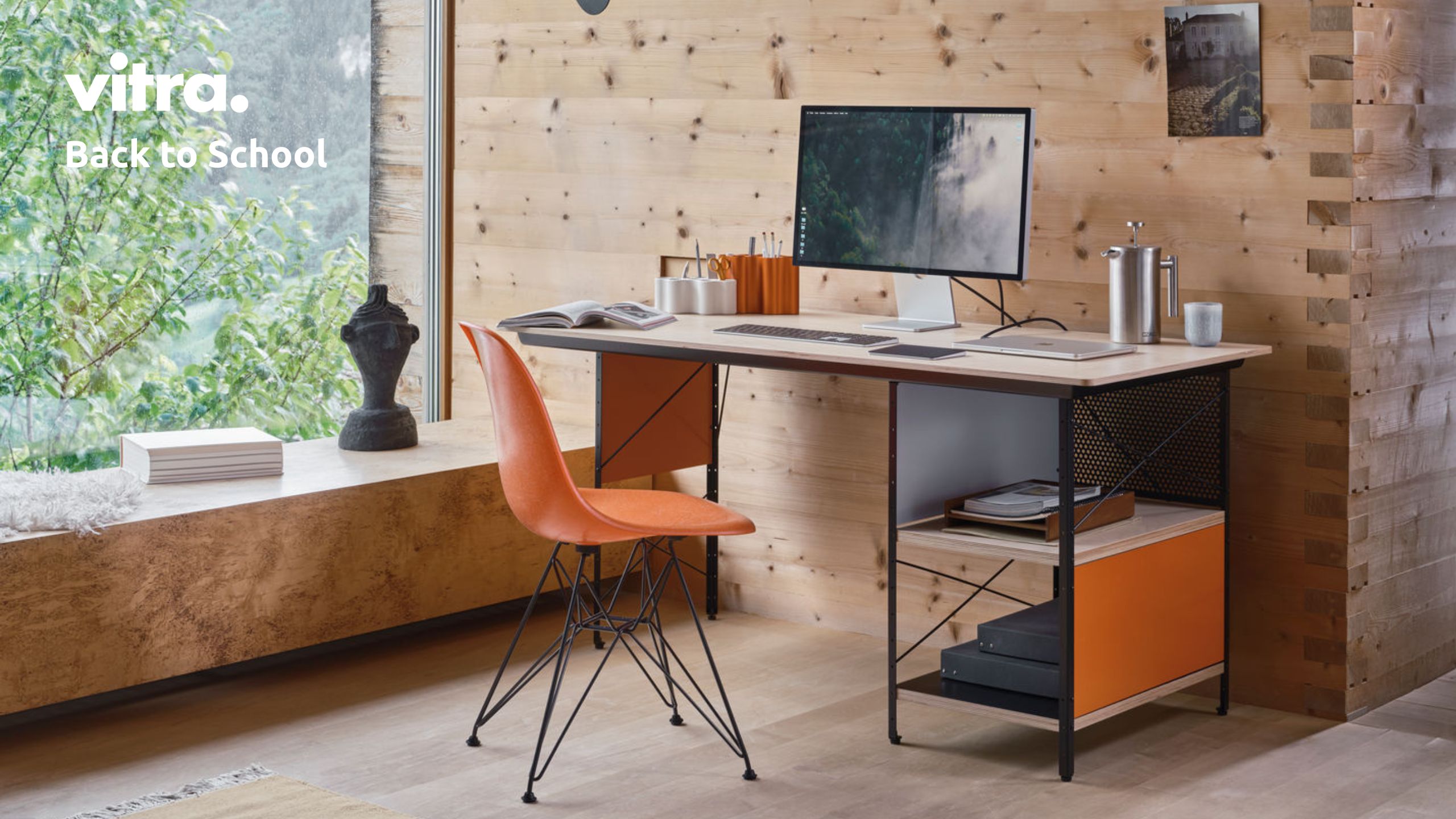 Ein modernes Heimbüro verfügt über einen Schreibtisch aus Holz mit orangefarbenen Akzenten, eine Eames-Schreibtischkombination, einen orangefarbenen „Back to School“-Stuhl von Vitra, einen Computermonitor, Bücher und Schreibwaren – vor dem Hintergrund einer holzvertäfelten Wand mit einem Fenster, das den Blick auf die grüne Umgebung freigibt.