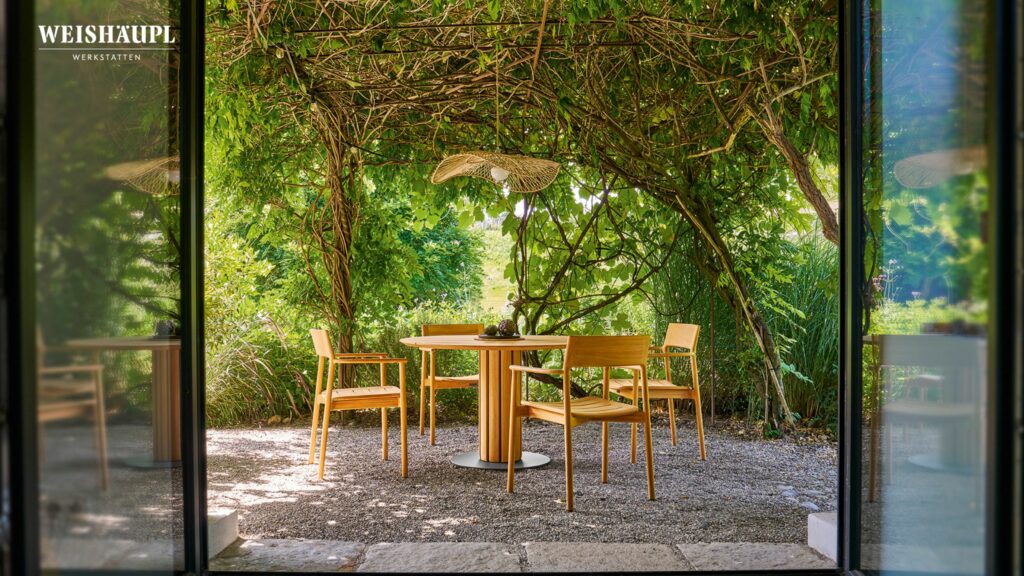 Outdoor-Sitzgruppe mit rundem Tisch und vier Holzstühlen unter einer begrünten Pergola, mit Blick durch eine offene Tür - perfekt für den Outdoor-Saisonstart. Das Logo WEISHÄUPL WERKSTÄTTEN erscheint in der oberen linken Ecke.