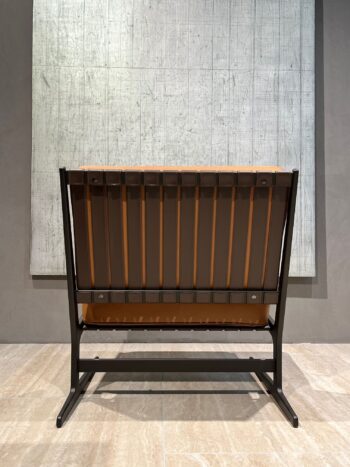 Ein brauner Paula-Sessel-Stuhl mit Metallrahmen steht von hinten gesehen auf einem Fliesenboden vor einem großen abstrakten grauen Wandkunstwerk - eine elegante Note, die an Molteni&C-Design erinnert.