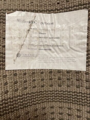 Ein Produktetikett für den Molteni&C Hedera Outdoor-Teppich mit Angaben zu Größe, Farbe, Material, Technik und Pflegeanleitung, platziert auf einer strukturierten Stoffoberfläche.