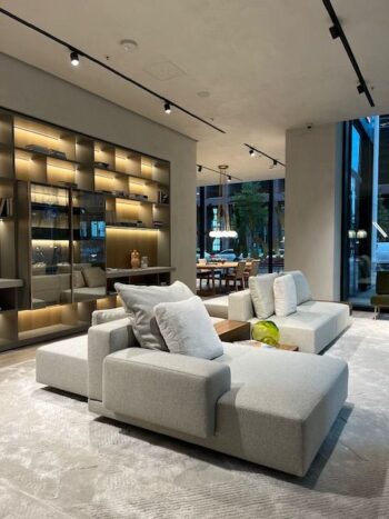 Modernes Wohnzimmer mit zwei hellgrauen Designersofas, eingebauten beleuchteten Regalen und großen Fenstern mit Blick auf einen Essbereich und Grünflächen im Freien. Das elegante Marteen Sofa von Molteni&C sorgt für einen Hauch von raffiniertem Komfort.