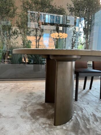 Ein Molteni&C Blevio Tisch mit Metallgestell steht auf einem hellen Teppich, umgeben von braunen Stühlen und hohen grünen Pflanzen, die durch eine Glaswand im Hintergrund sichtbar sind.