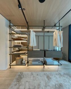 Moderner begehbarer Kleiderschrank mit offenen Regalen, hängenden Kleidungsstücken, Aufbewahrungsboxen, gefalteten Handtüchern, einem Korb und einem Design aus hellem Holz und Metall von Molteni&C Ankleide. Im Vordergrund ist der Teppichboden zu sehen.