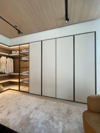 Moderner begehbarer Kleiderschrank von Molteni&C mit beigefarbenen Schiebetüren von Gliss Master, offenen Regalen mit gefalteter Kleidung und hängenden weißen Kleidungsstücken - alles wunderschön beleuchtet von einer Einbauleuchte.