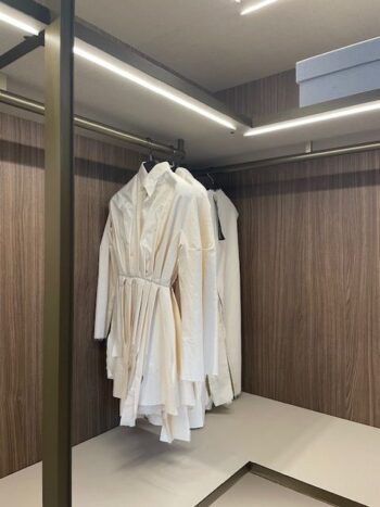 Vier weiße Kleidungsstücke, darunter ein langärmeliges Kleid, hängen auf einer Stange in einem modernen Molteni&C Gliss Master-Schrank mit holzgetäfelten Wänden und Deckenbeleuchtung.