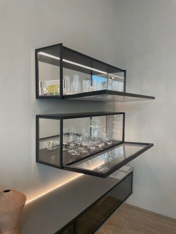 Drei schwarz gerahmte Glasregale aus der Serie Molteni&C Pass-Word Evolution sind an einer Wand montiert, zwei davon mit offenen Türen und bestückt mit Klarglas. Unter diesem stilvollen Wandmodul ruht ein Holzobjekt auf der Theke.
