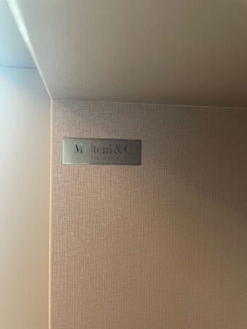 Eine Metallplakette an einer strukturierten beigen Wand trägt den Markennamen Molteni&C und zeigt das Pass-Word Evolution Wandmodul, darunter steht Made in Italy.