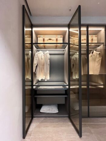 Der moderne begehbare Kleiderschrank Gliss Master von Molteni&C verfügt über Glastüren, in denen weiße Kleidung, Gepäckstücke auf dem obersten Regal und ein Kissen auf dem untersten Regal bei gedämpfter Beleuchtung präsentiert werden, um einen eleganten Kleiderschrank zu schaffen.