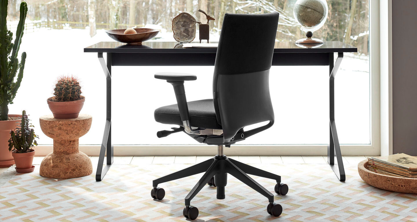 Schwarzer Bürostuhl an einem modernen Schreibtisch mit Dekorationsgegenständen, vor einem großen Fenster mit Blick auf die verschneite Landschaft; in der Nähe stehen mehrere Topfpflanzen auf dem Boden.