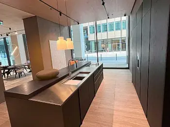 Molteni Abverkauf, Outlet, Angebot: Moderne Küche mit dunklen Schränken, Molteni&C-Insel mit eingebauter Spüle, Pendelleuchten und großen, raumhohen Fenstern mit Blick auf eine Stadtstraße. Das Design von Ratio Essenza Küche hebt hochwertige Küchengeräte hervor.